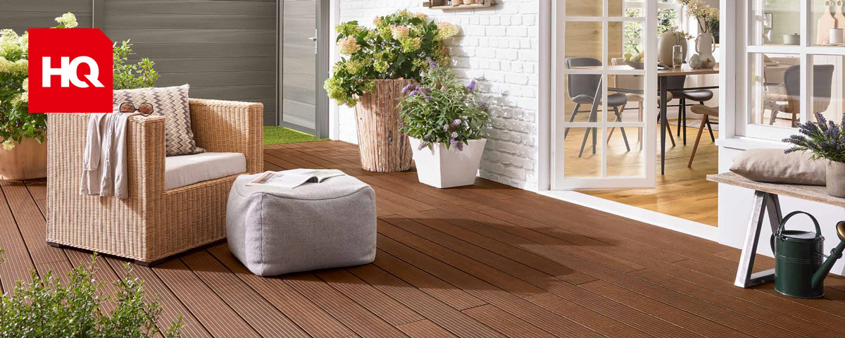 Holzfarbene Terrasse mit einem Flechtsessel und Pouf sowie bepflanzten Kübeln vor einer weißen Hauswand.